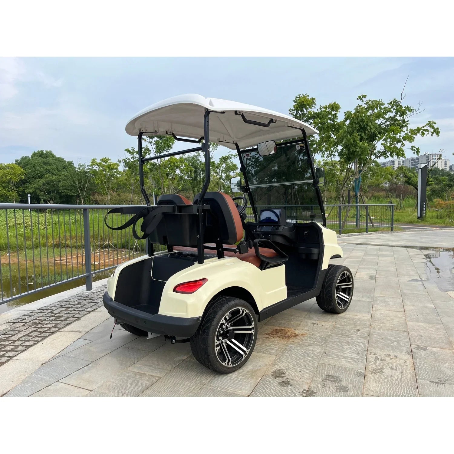 UAE golf cartz rear three-quarter view of a cream-colored four-seater luxury golf cart with a white roof, parked on a paved path overlooking a body of water in a park setting. This electric vehicle features brown and black seats, a rear cargo area with a bag attachment, and a tinted windshield. The cart is equipped with low-profile tires and custom black and silver alloy wheels, highlighting its sleek NEV or resort transport design.
