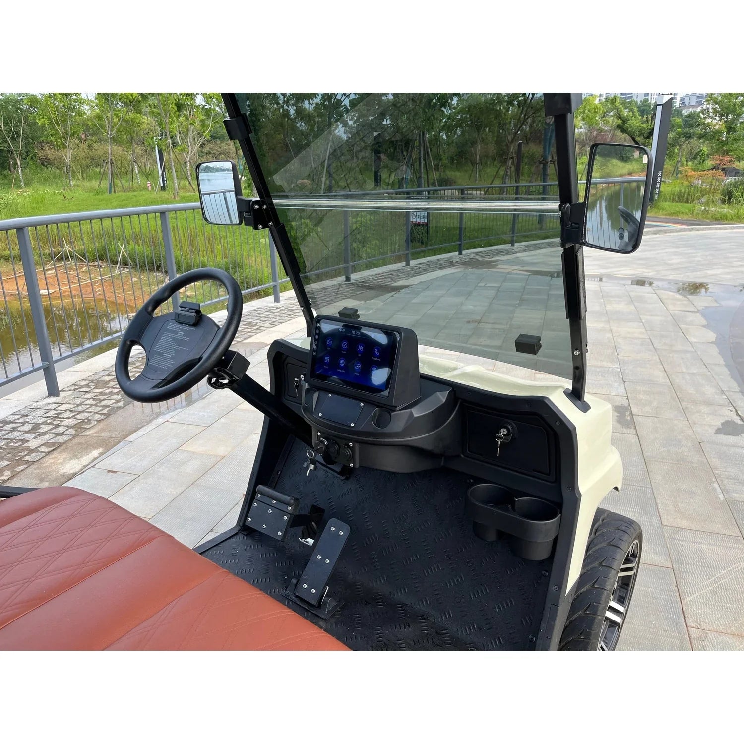 UAE golf cartz close-up interior view of a cream-colored luxury golf cart cockpit, focusing on the steering wheel and dashboard. The image highlights the digital touchscreen display mounted centrally, a black steering wheel, two cup holders, and textured black floor mats. The driver's area shows a brown seat edge. The electric vehicle is parked outdoors on a paved area overlooking a pond or waterway, emphasizing its features as a premium NEV or resort transport cart.