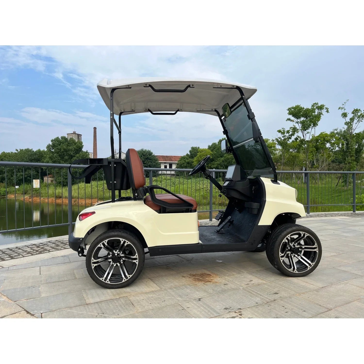 UAE golf cartz side profile view of a cream-colored four-seater luxury golf cart with a white roof, parked on a paved stone path next to a dark body of water. This electric vehicle features brown and black seats, a modern front end, and a tinted windshield. The lifted golf cart is equipped with low-profile tires and custom black and silver alloy wheels, set against a partially industrial background with trees and a metal railing, highlighting its stylish NEV or resort transport design.