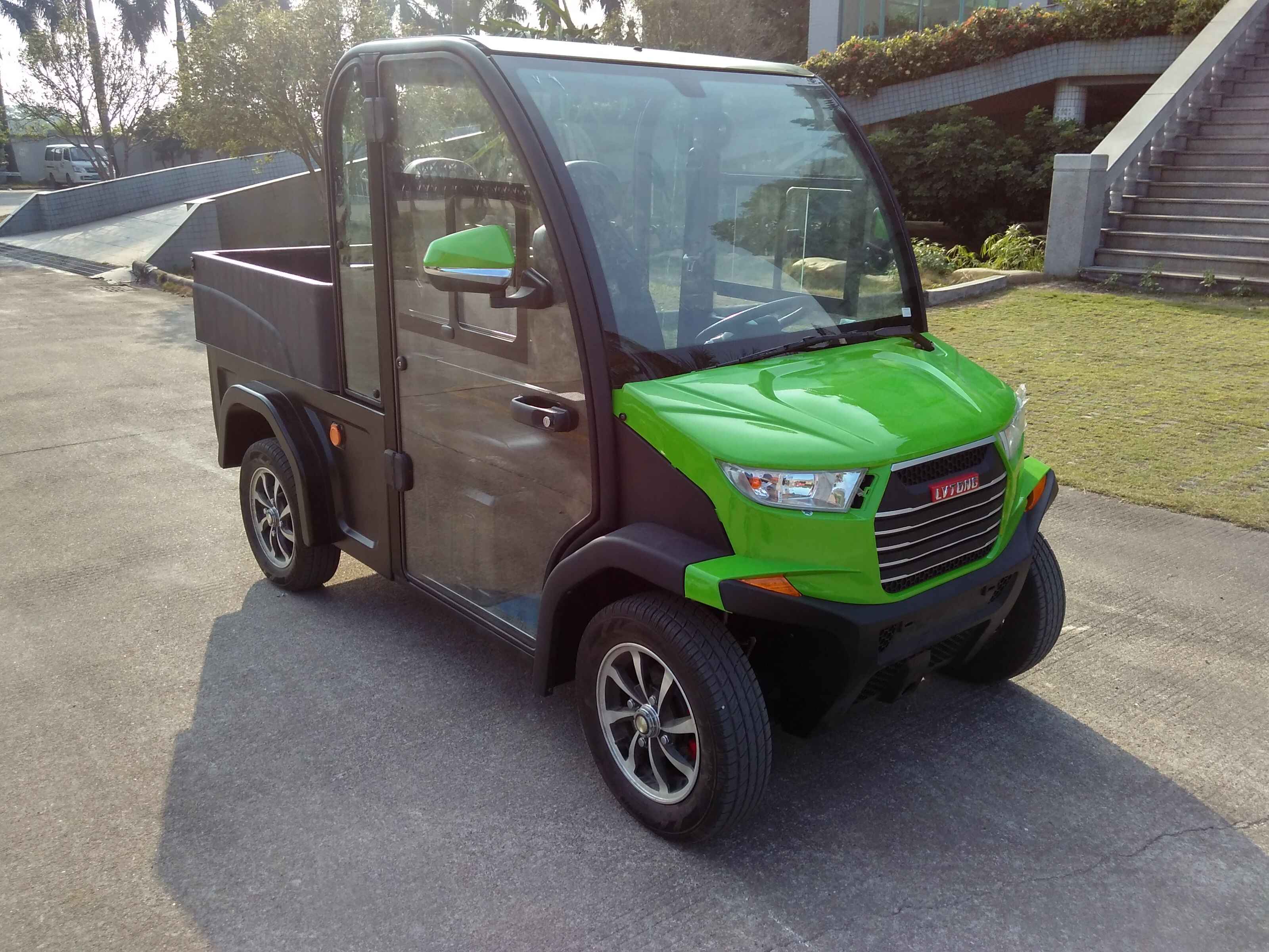 UAE golf cartz three-quarter front view of a lime green and black utility electric vehicle with a fully enclosed cab and a black rear cargo bed. This two-seater commercial NEV or electric work cart features a bright green hood, black enclosed doors with glass, and a large front windshield. It is equipped with custom black and silver wheels and is parked on an asphalt and concrete area outside a modern building with steps and landscaping, highlighting its use as a compact utility transport vehicle.