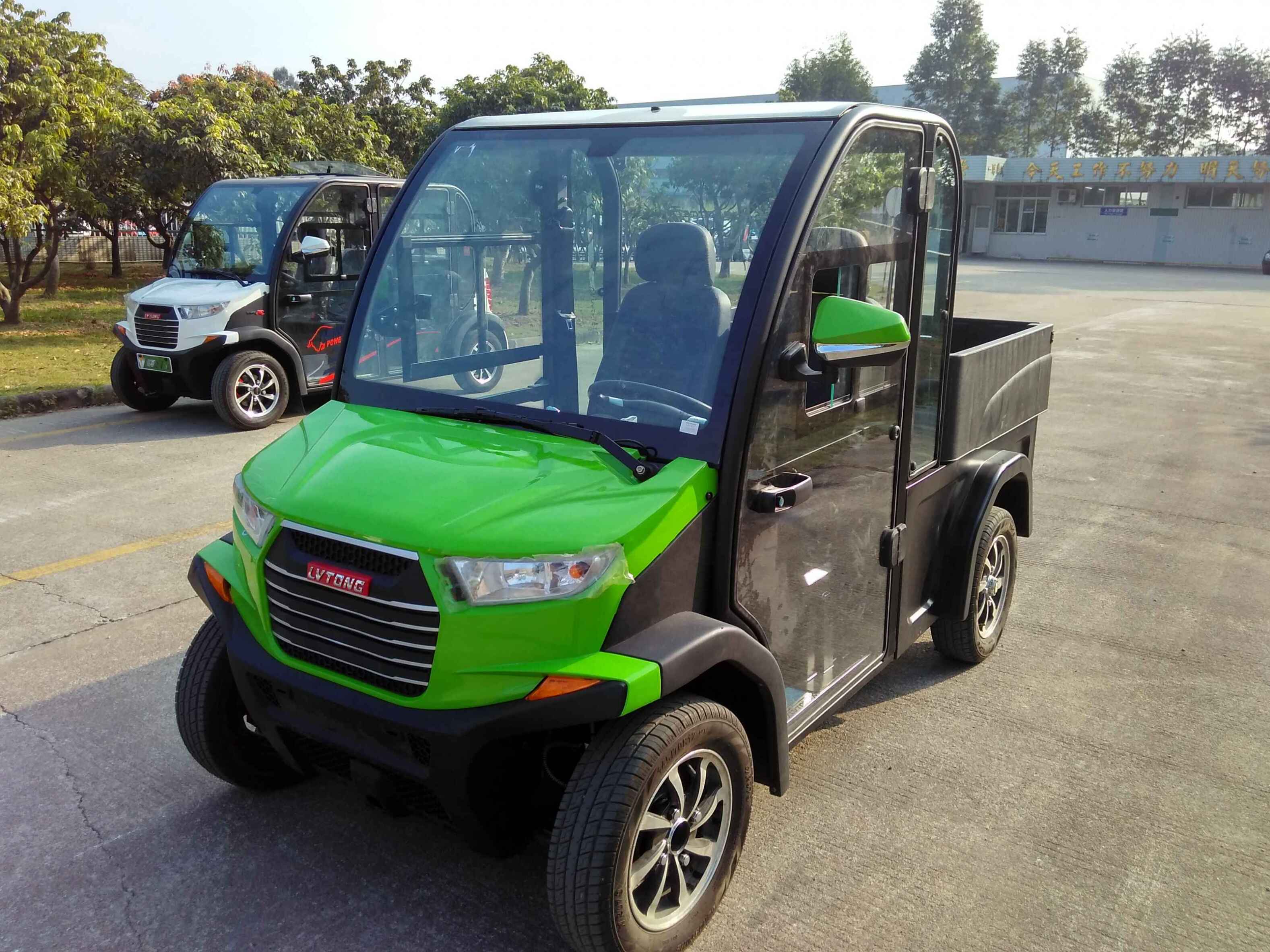 UAE golf cartz three-quarter front view of a lime green and black utility electric vehicle with a fully enclosed cab and a black rear cargo bed, parked on an asphalt lot. This two-seater commercial NEV or electric work cart features a bright green hood, black enclosed doors with glass, and a modern front grille. Another similar vehicle is parked in the background, suggesting a fleet. The utility transport vehicle is outdoors with trees and buildings in the distance.