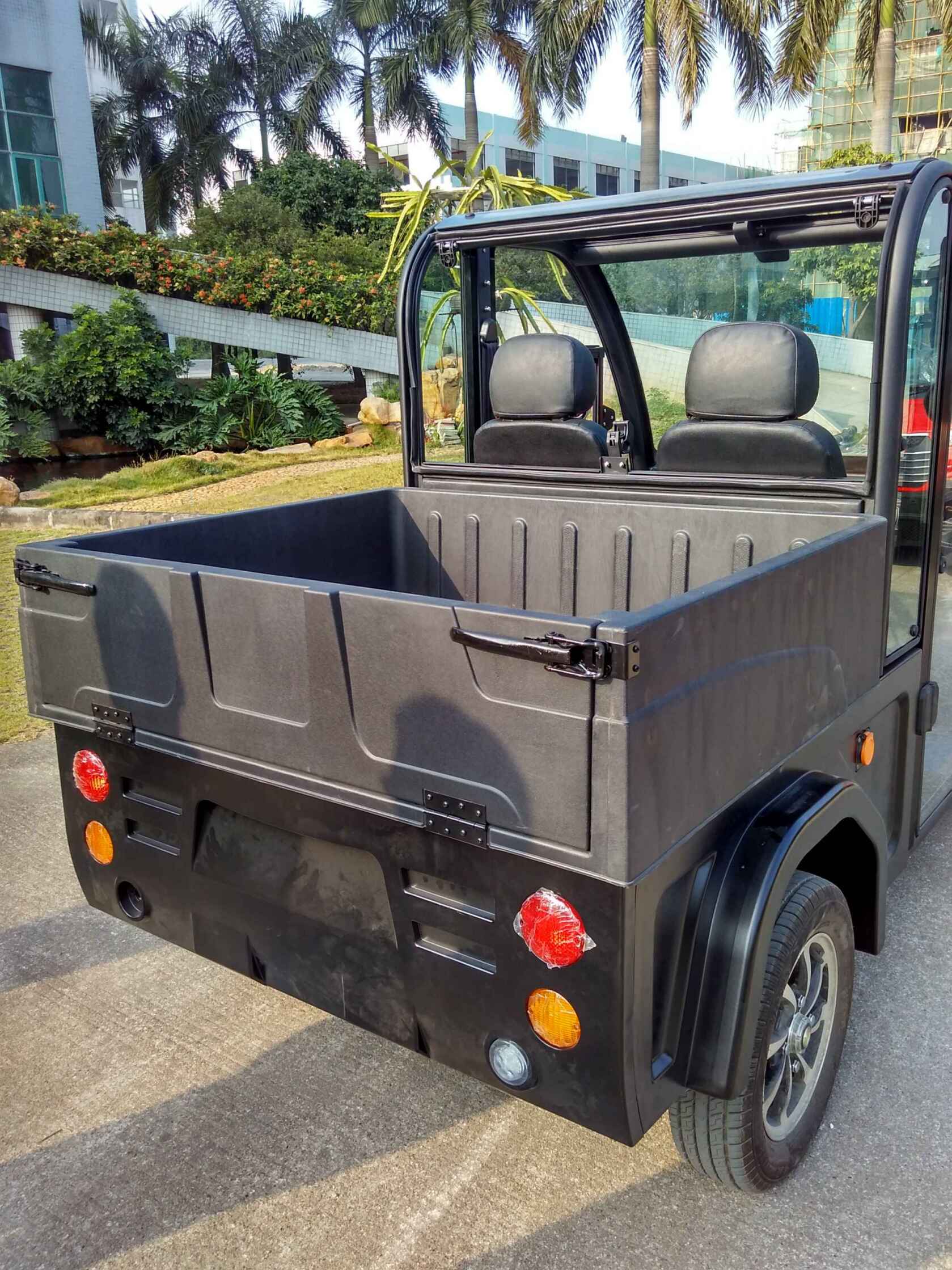 UAE golf cartz rear view of a black electric utility vehicle with a durable, molded plastic cargo box and an open-top cabin, parked outdoors. This two-seater electric work cart features black seats, a clear rear window, and round red and amber taillights. The image highlights the deep hauling capacity of the utility transport vehicle, set in a sunny area with palm trees and a modern building in the background.