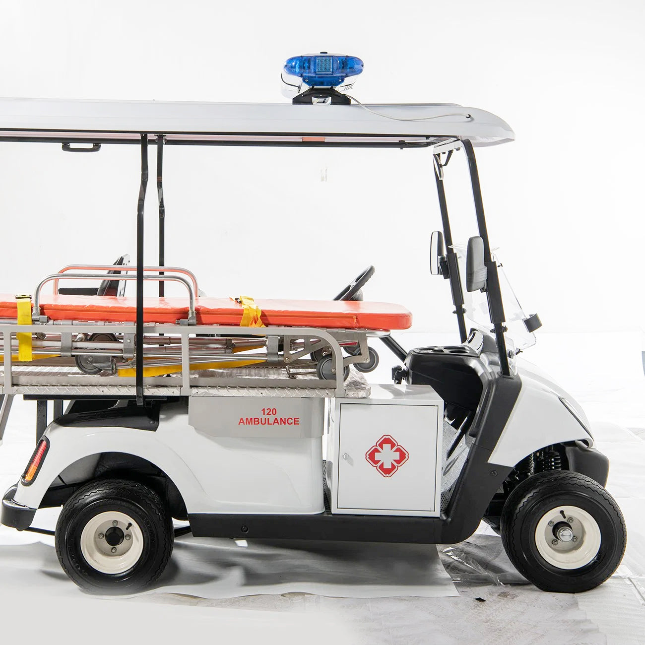 UAE golf cartz side view studio shot of a white electric ambulance golf cart or medical utility vehicle against a white background. This specialized emergency response cart features a stretcher with an orange pad and yellow straps mounted in the rear. It has a blue emergency light on the roof, a side storage cabinet marked with a red cross logo, and a side panel marked "120 AMBULANCE", clearly indicating its function as a rescue golf cart.