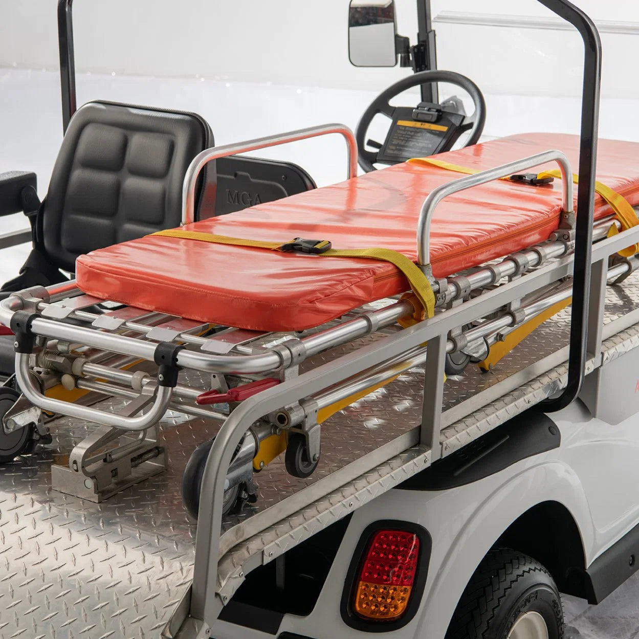 UAE golf cartz close-up interior view of a white ambulance golf cart focusing on the patient transport area. The image clearly shows a metallic, diamond-plated rear deck holding a silver emergency stretcher with an orange pad and yellow safety straps. The driver's black seat and steering wheel are visible in the background, highlighting the specialized equipment of this rescue golf cart or medical utility vehicle.