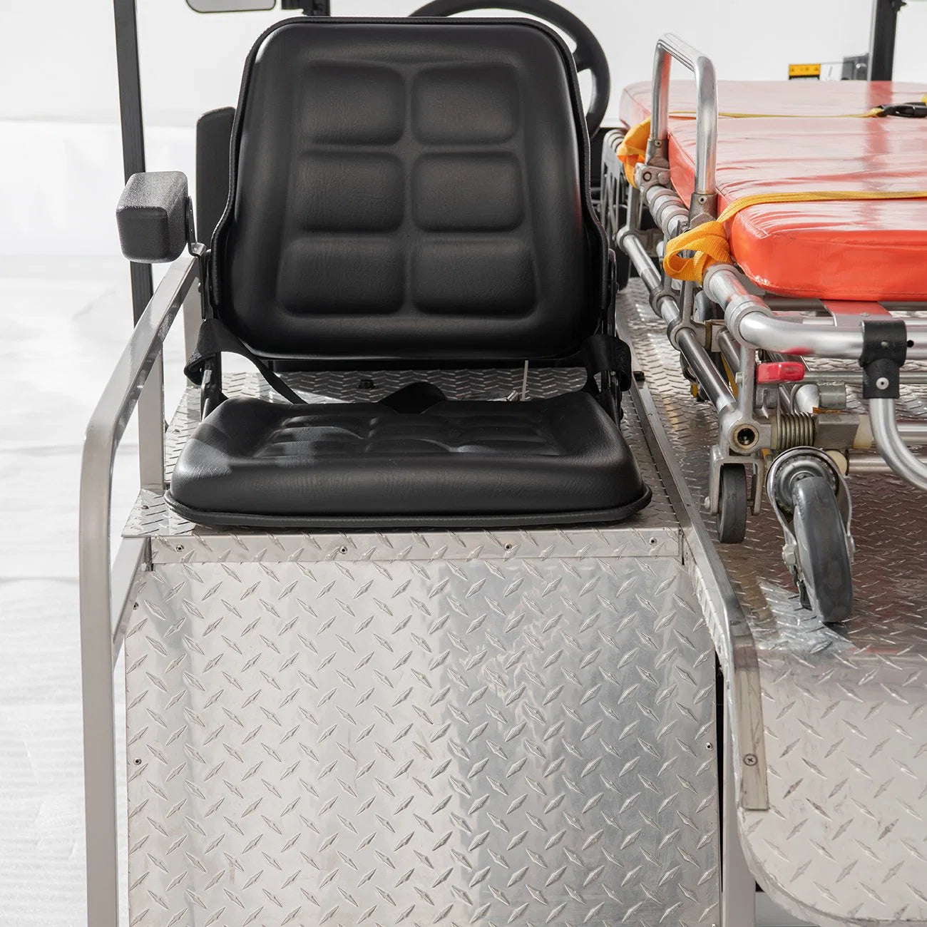 UAE golf cartz close-up detail of the black driver's seat and the diamond-plate metal deck inside a white ambulance golf cart. The utility cart seat is a wide, black, quilted design with armrests. To the right, the rear section of the cart shows the wheels and frame of the orange emergency stretcher, highlighting the specialized, durable construction of the medical utility vehicle.