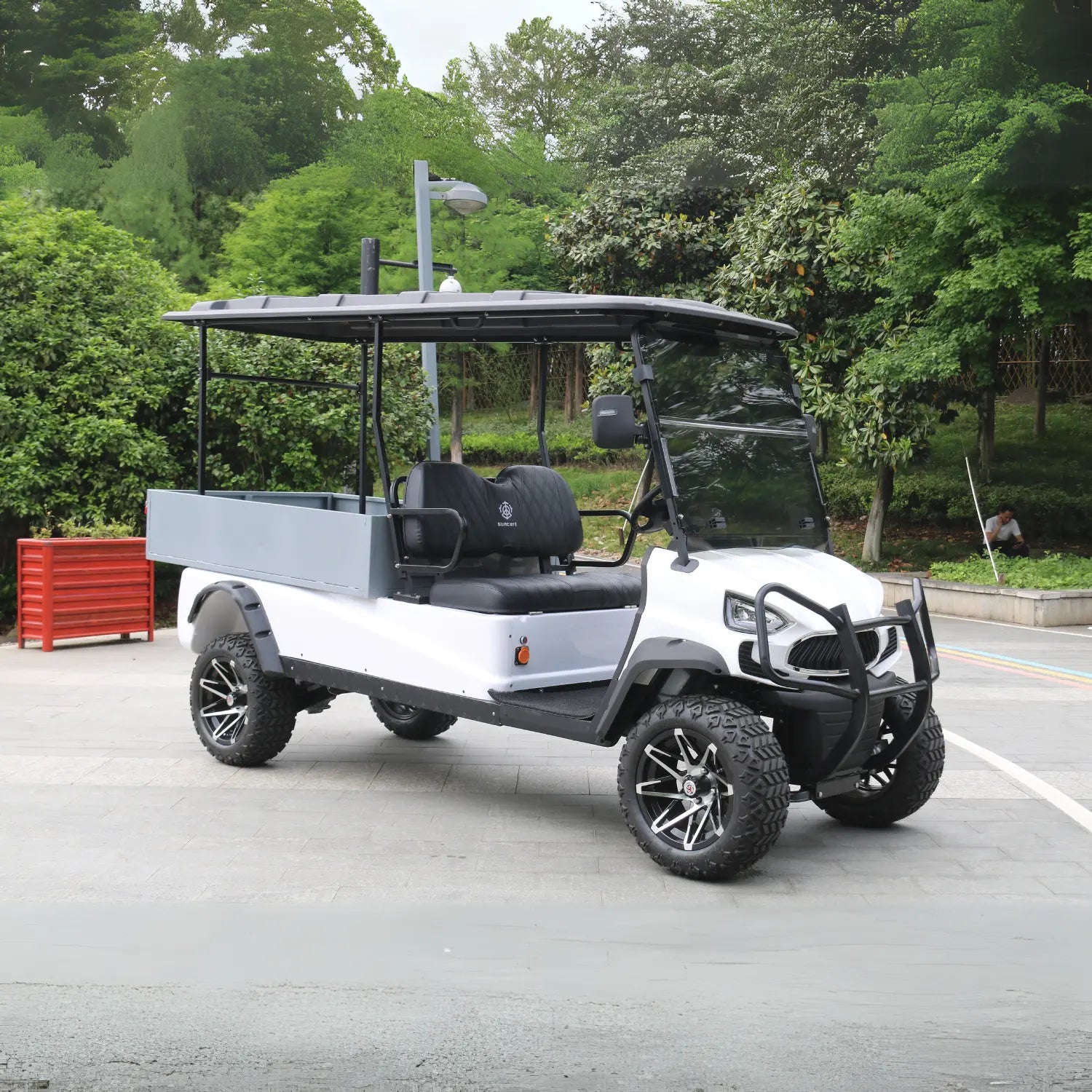 UAE golf cartz side view of a white utility golf cart with a large, grey rear cargo box and a black roof, parked on an asphalt path. This lifted electric vehicle is an off-road utility cart featuring black quilted seats, a front brush guard, and aggressive all-terrain tires. The wheels are custom black and silver alloy designs, and the vehicle is shown in a park or campus setting surrounded by dense green trees.