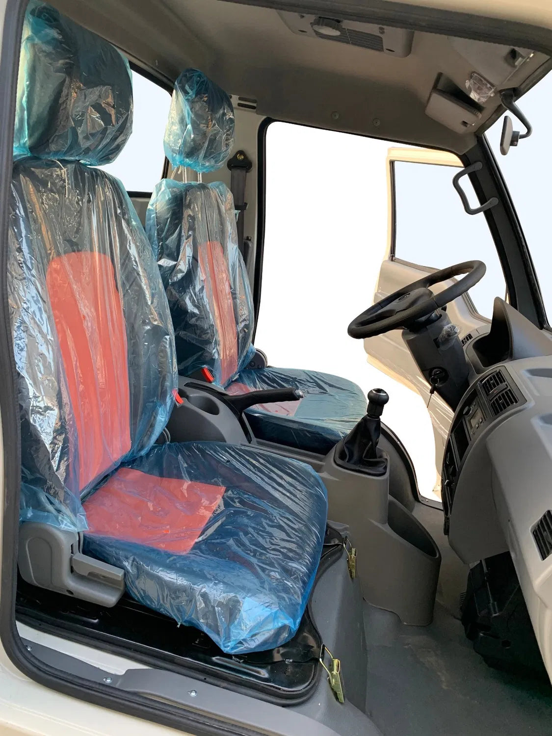 UAE golf cartz close-up interior view of the two-seater cabin in a white electric box truck or delivery van, seen from the open driver's door. The seats feature red and black upholstery covered in clear plastic for protection. The image prominently shows the black steering wheel, manual gear shifter, and hand brake, highlighting the compact and functional design of the electric commercial utility vehicle.