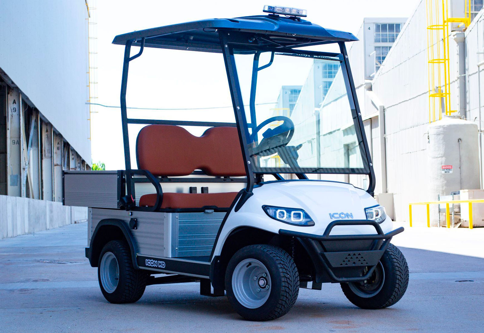 UAE golf cartz three-quarter front view of a white utility electric vehicle with a black roof and a silver aluminum rear cargo bed, parked in an industrial setting. This specialized two-seater work cart features brown seats, a protective front brush guard, a large windshield, and a light bar on the roof. The electric utility golf cart is equipped with silver utility wheels and all-terrain tires, highlighting its heavy-duty hauling and property maintenance function.