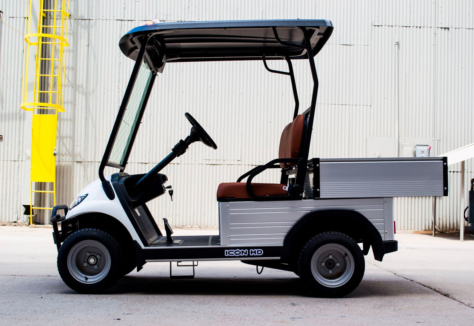 UAE golf cartz side profile view of a white utility electric vehicle with a black roof and a silver aluminum rear cargo bed with drop-down sides. This specialized two-seater electric work cart features brown seats, a steering wheel, and silver utility wheels. The electric utility golf cart is parked on a paved surface next to a corrugated metal industrial building, with a yellow ladder visible in the background, highlighting its heavy-duty property maintenance function.