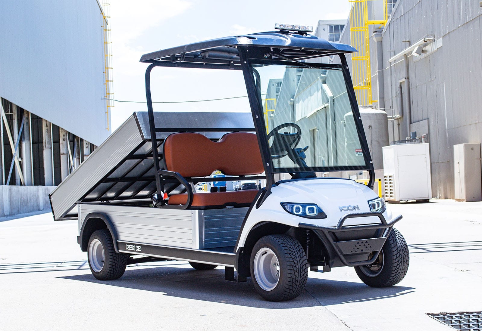 UAE golf cartz three-quarter front view of a white utility electric vehicle with a fully electric dump bed raised for unloading, parked in an industrial area. This specialized electric work cart features a black roof, a black brush guard, and brown two-seater upholstery. The utility golf cart has custom wheels and a clear windshield, with the large aluminum dump bed prominently displayed.