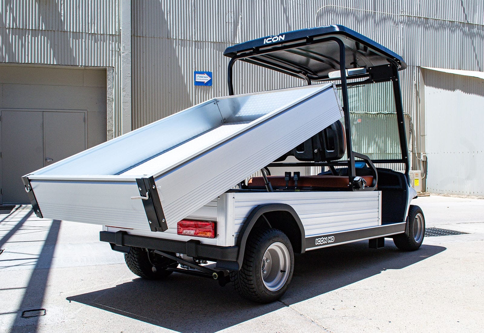 UAE golf cartz rear three-quarter view of a white utility electric vehicle with a fully raised dump bed, demonstrating its unloading capability. This specialized electric work cart features a silver aluminum cargo bed, black roof, and a low profile. The dump cart is parked in an industrial area, highlighting its heavy-duty design for commercial hauling and utility operations.