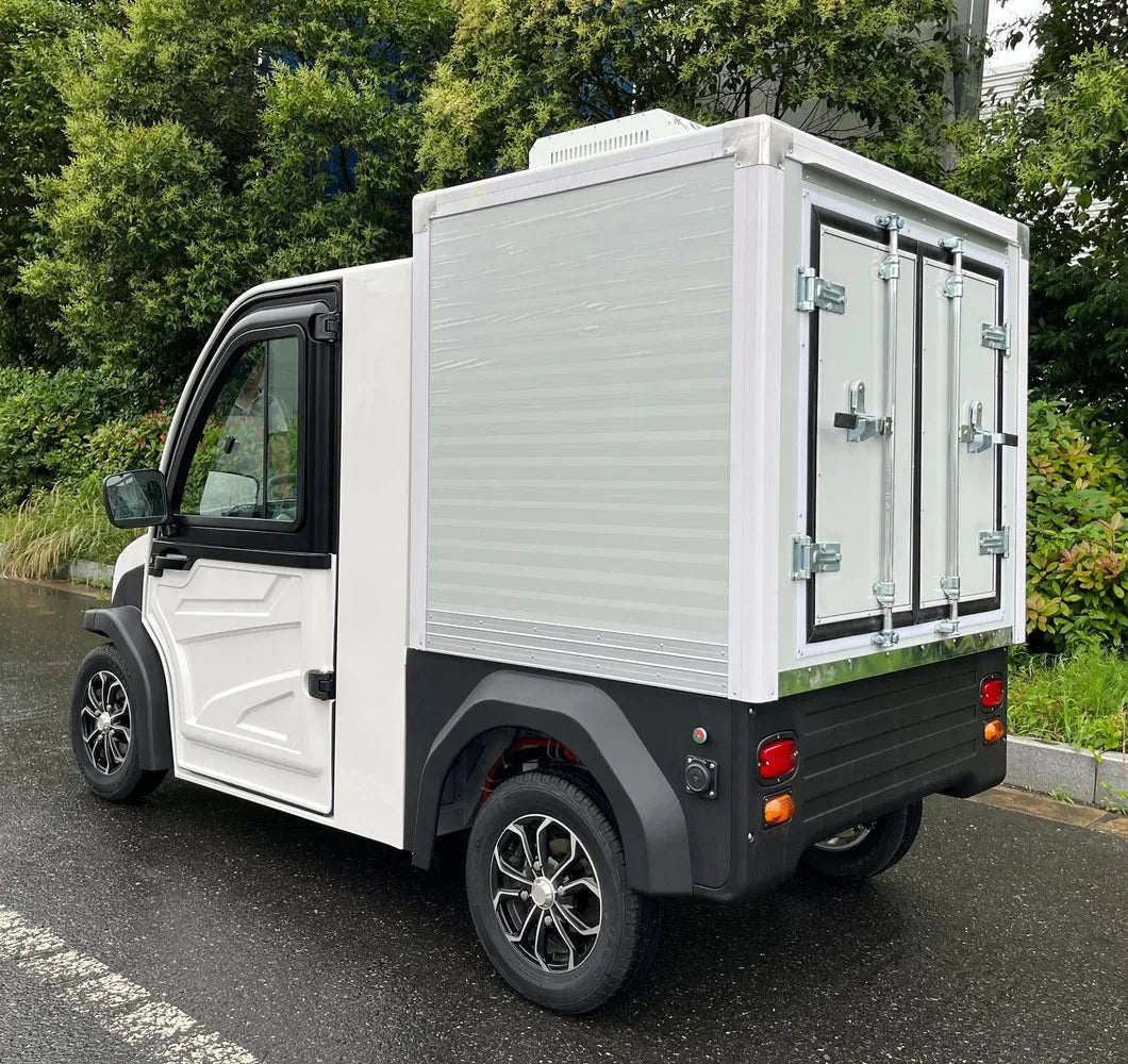 UAE golf cartz rear three-quarter view of a white electric refrigerated delivery vehicle with an enclosed cab, parked on a wet asphalt road. This compact electric NEV or refrigerator work cart features a silver insulated rear cargo box with double doors and a top-mounted refrigeration unit. The vehicle has black trim, black fender flares, and custom black and silver wheels, highlighting its use for cold chain last-mile delivery.