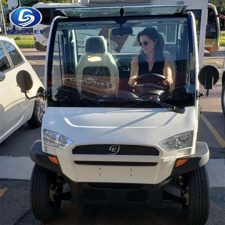 UAE golf cartz head-on view of a white two-seater electric vehicle with a fully enclosed cab. A woman wearing sunglasses is sitting in the driver's seat. This compact NEV or low-speed electric car features a modern front end, a small grille, and a clear windshield. The image is taken outdoors in a parking lot, with other vehicles and a bus visible in the background, showing the electric vehicle in use.