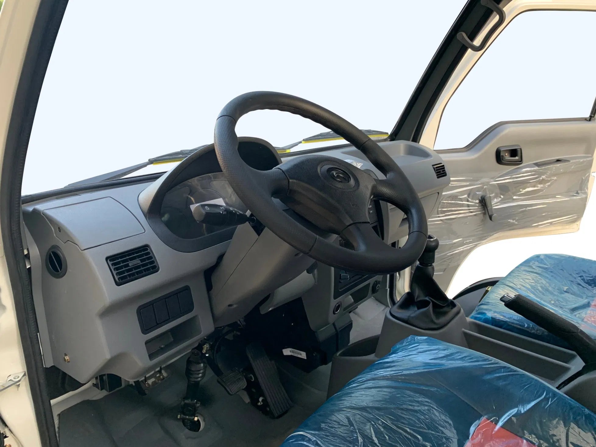 UAE golf cartz close-up interior view of the driver's cabin in a white electric flatbed truck. The image highlights the grey dashboard, black four-spoke steering wheel, instrument panel, air vents, and manual gear shift lever. The seats are covered in protective plastic, showcasing the simple and functional design of the electric commercial utility vehicle or work truck.