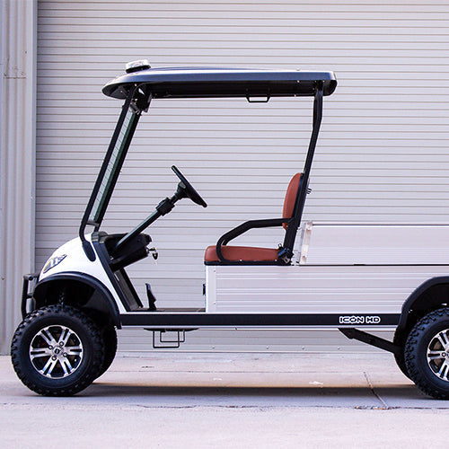 UAE golf cartz side profile view of a white utility electric vehicle with a black roof and a silver aluminum rear cargo bed, parked in front of a light gray roll-up garage door. This specialized two-seater work cart features brown seats and a lifted suspension. The electric utility golf cart is equipped with all-terrain tires and custom black and silver wheels, highlighting its heavy-duty property maintenance function.
