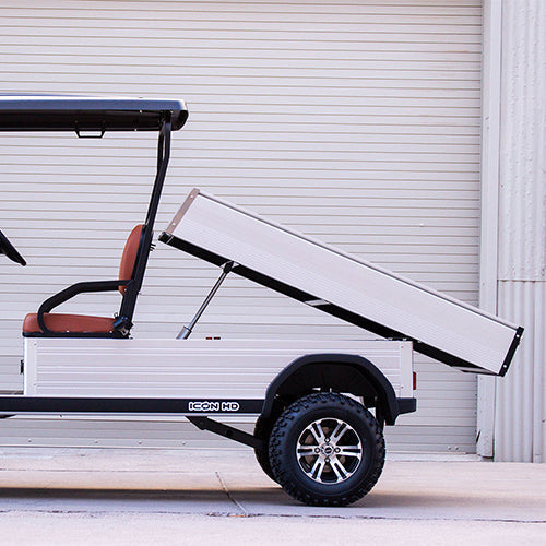 UAE golf cartz side profile close-up of a white utility electric vehicle demonstrating its fully raised dump bed function, parked in front of an industrial roll-up door. This specialized two-seater electric work cart features brown seats and a silver aluminum rear cargo bed with a hydraulic lifting mechanism clearly visible. The lifted utility golf cart is equipped with all-terrain tires on custom black and silver wheels, highlighting its heavy-duty hauling and dumping capability.
