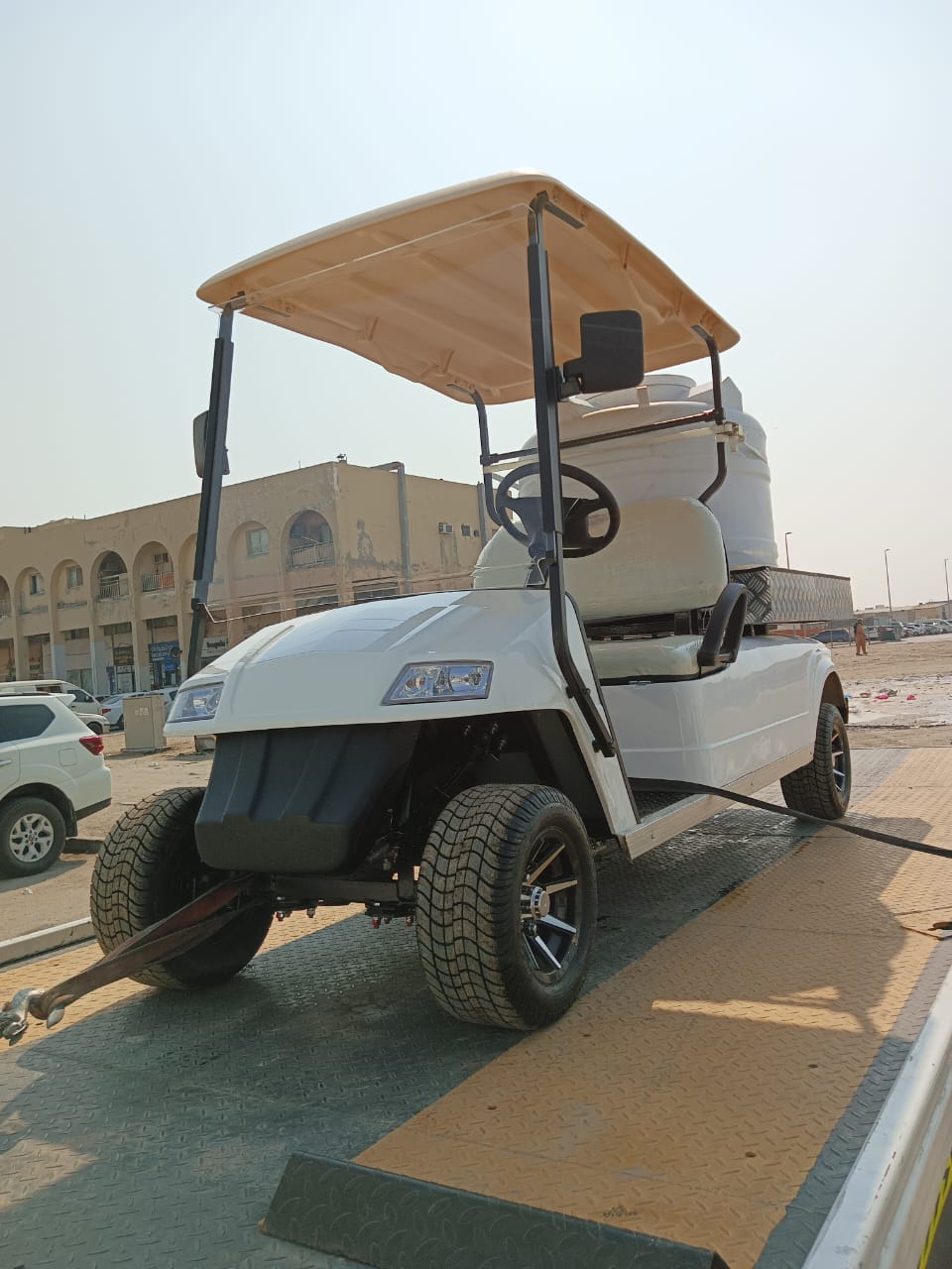 UAE golf cart three-quarter front view of a white utility electric vehicle designed as a sprayer or water transport golf cart, secured on the flatbed of a tow truck or trailer. This specialized two-seater work cart features a large white spherical water tank mounted in the rear and a beige roof. The lifted cart is equipped with all-terrain tires and custom black wheels, highlighting its function as a maintenance vehicle being transported in an urban or industrial area.