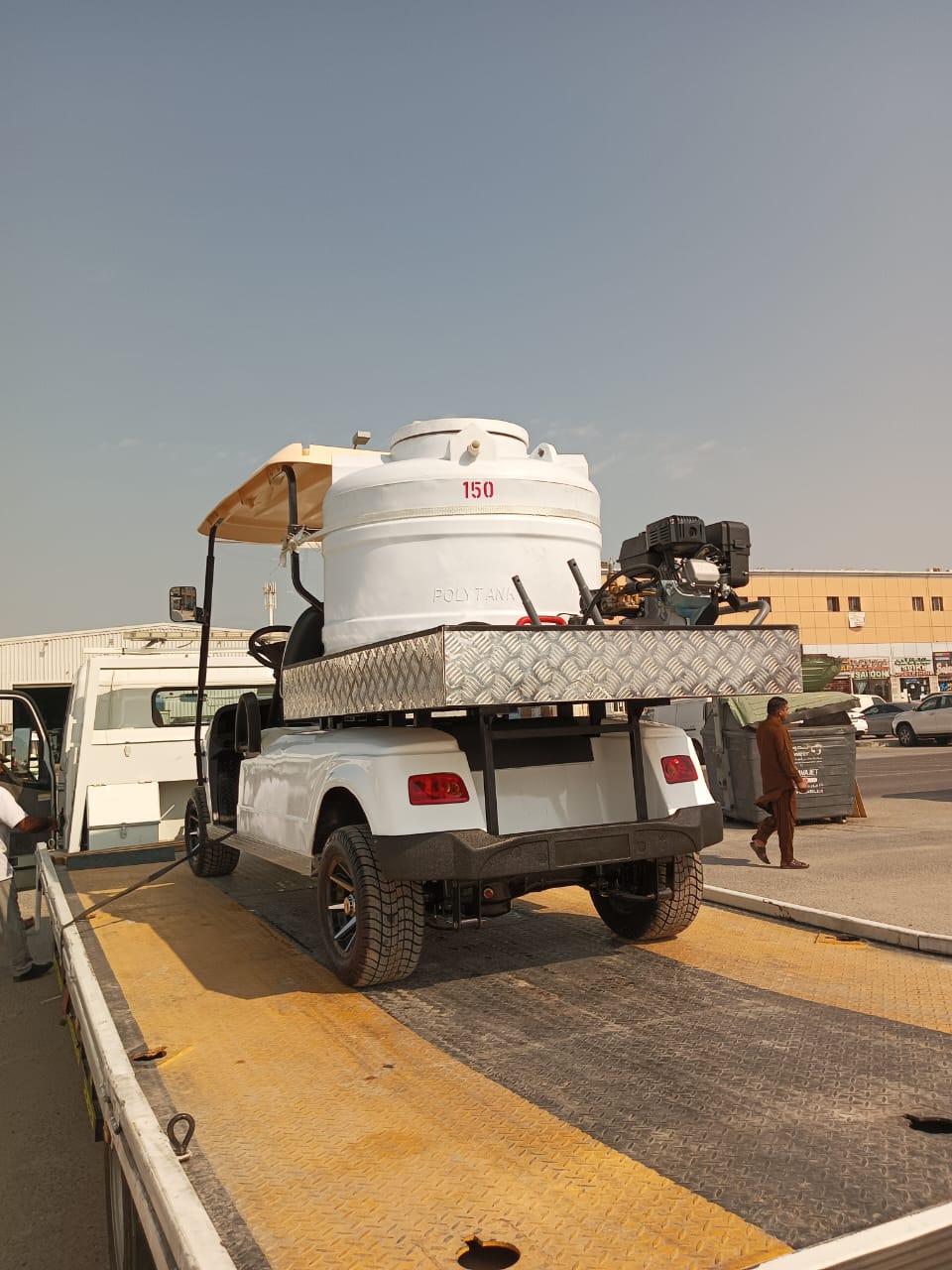 UAE golf cartz rear three-quarter view of a white utility electric vehicle designed as a sprayer or water transport golf cart, being loaded onto a flatbed tow truck or trailer. This specialized two-seater work cart features a large white cylindrical water tank labeled "150" and "POLY TANK," mounted on a metal diamond-plate platform with a gas-powered pump or engine. The lifted utility cart has a beige roof, all-terrain tires, and custom black wheels, shown in a sunny industrial or commercial area.