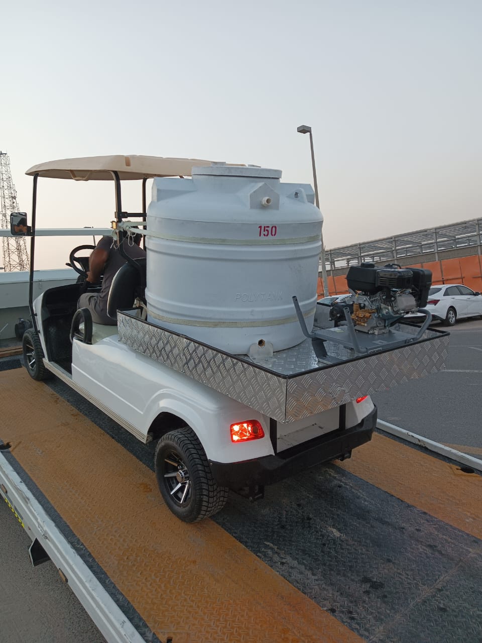 UAE golf cartz rear three-quarter view of a white utility electric vehicle designed as a sprayer or water transport golf cart, being loaded or unloaded from a flatbed tow truck or trailer. This specialized two-seater work cart features a large white cylindrical water tank labeled "150" and "POLY TANK," mounted on a metallic diamond-plate platform with a gas-powered pump or engine. The lifted utility cart has a beige roof and custom black and silver wheels, shown in a sunny industrial area.