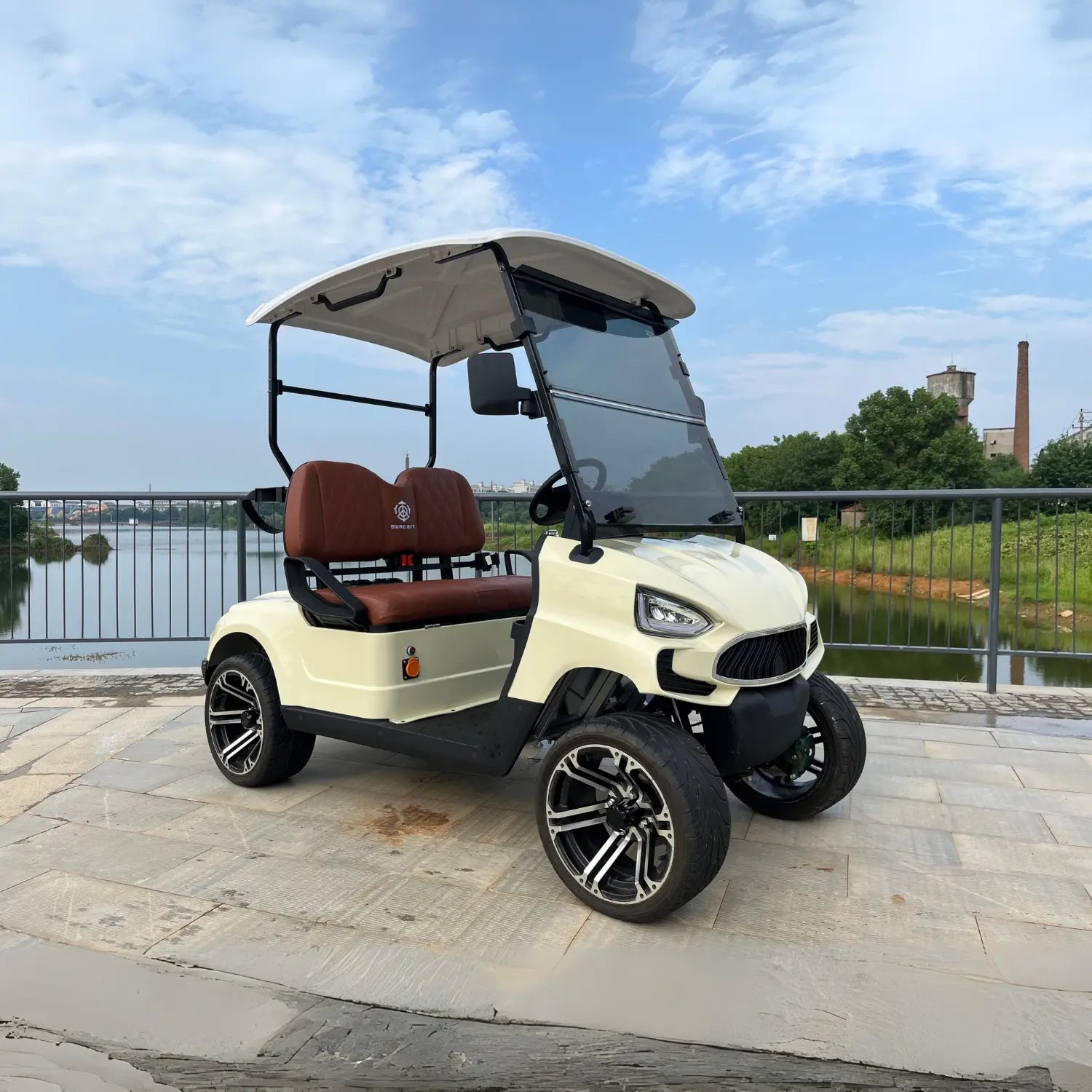 UAE golf cartz three-quarter front view of a cream-colored four-seater luxury golf cart with a white roof, parked on a paved walkway next to a body of water. This electric vehicle features brown leather seats with diamond stitching, a modern front end, and a tinted windshield. The lifted golf cart is equipped with all-terrain tires and custom black and silver alloy wheels, highlighting its stylish and premium resort or NEV design.