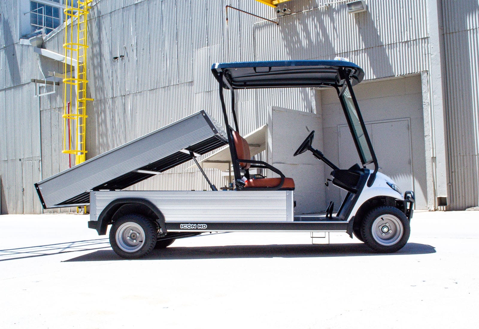 UAE golf cartz side profile view of a white utility electric vehicle with a fully raised silver aluminum dump bed, demonstrating the hydraulic function. This specialized electric work cart features a black roof and a single row of brown seating. The dump cart is parked in front of a corrugated metal industrial building, highlighting its use as a heavy-duty hauling utility vehicle.