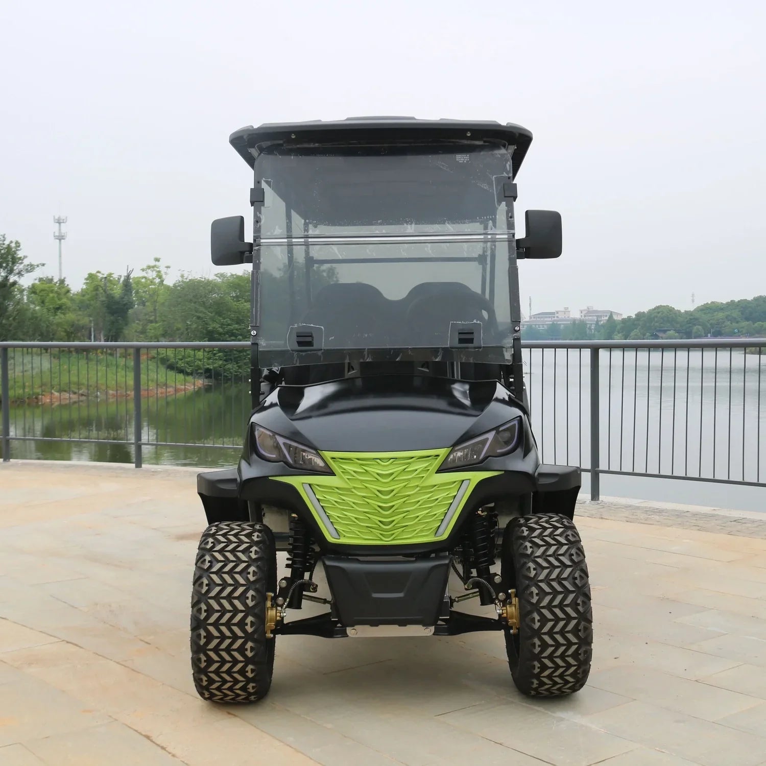 Green and black off-road vehicle on a paved area with a lake and trees in the background