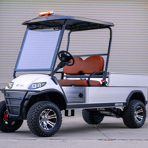 UAE golf cartz three-quarter front view of a white utility electric vehicle with a black roof and a silver aluminum rear cargo bed, parked in an industrial setting. This specialized two-seater work cart features brown seats, a black brush guard, a large front windshield with a solar panel on it, and an amber emergency light bar on the roof. The lifted utility golf cart is equipped with knobby all-terrain tires on custom black and silver wheels, highlighting its heavy-duty property maintenance function.